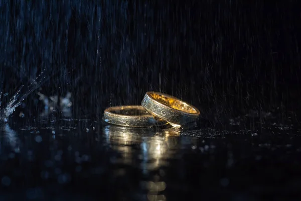 Anillos de boda que yacen en la superficie oscura brillando con luz. Salpicaduras de agua — Foto de Stock