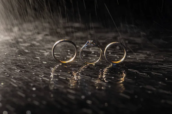 Anéis de casamento deitado na superfície escura brilhando com luz. Salpicos de água. Chuva — Fotografia de Stock