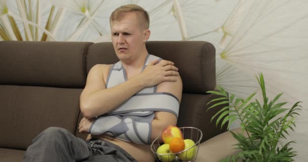 Schmerzhafter Mann mit gebrochenem Arm und Armspange, der auf einem Sofa sitzt und Apfel isst — Stockvideo