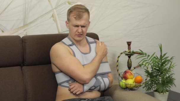 Painful, bored man with a broken arm wearing arm brace sitting on a sofa — Stock Video