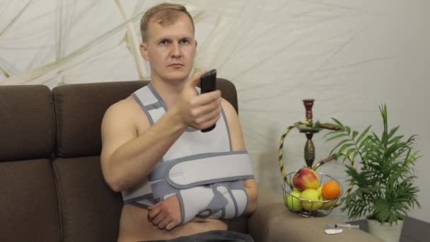Doloroso hombre con un brazo roto usando un brazalete sentado en un sofá viendo la televisión — Vídeo de stock