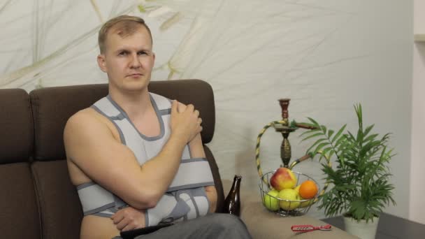 Homem doloroso com um braço quebrado usando cinta de braço sentado em um sofá assistindo TV — Vídeo de Stock
