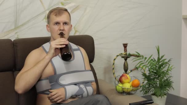 Pijnlijke man met een gebroken arm dragen arm brace zittend op een bank drinken bier — Stockvideo