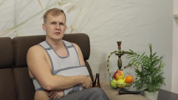 Doloroso hombre con un brazo roto usando un brazalete sentado en un sofá bebe cerveza — Vídeos de Stock