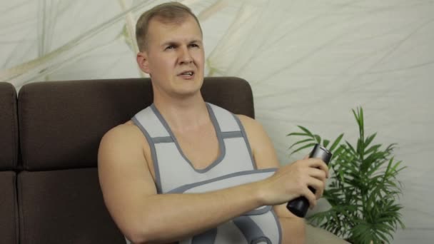 Homem doloroso com um braço quebrado usando cinta de braço sentado em um sofá assistindo TV — Vídeo de Stock