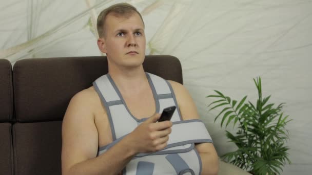 Больной мужчина со сломанной рукой в корсете для рук сидит на диване и смотрит телевизор. — стоковое видео