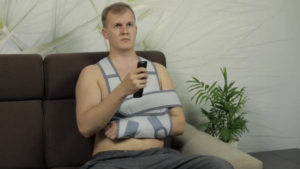 Pijnlijke man met een gebroken arm dragen arm brace zittend op een bank kijken tv — Stockvideo