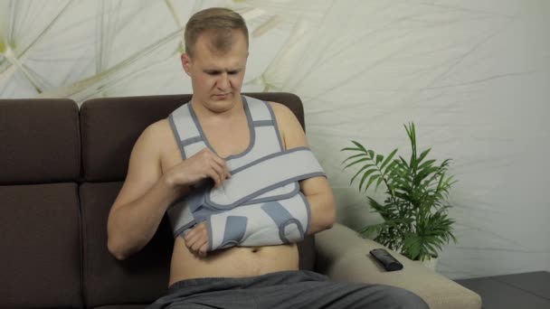 Bolestivé, znuděný muž se zlomenou rukou nošení paži sedí na pohovce — Stock video