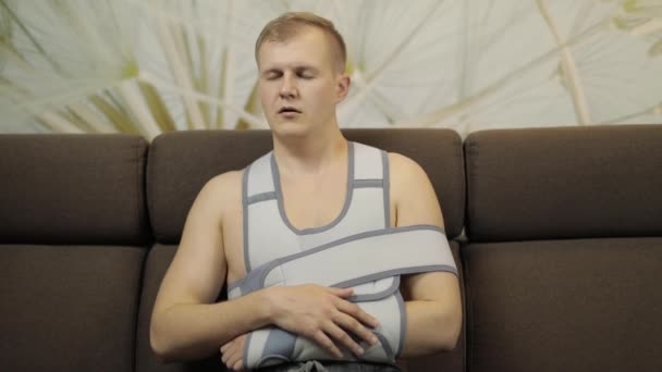 Pijnlijke, verveeld man met een gebroken arm dragen arm brace zittend op een bank — Stockvideo