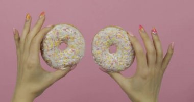 Kadınların elleri pembe arka planda iki beyaz, lezzetli, serpiştirilmiş donut tutuyor..