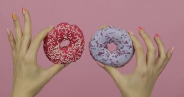 Kadınların elleri pembe arka planda iki kırmızı ve mavi, serpiştirilmiş donut tutuyor..