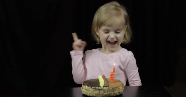 Маленькая милая девочка задувает праздничную свечу на торте на черном фоне — стоковое видео