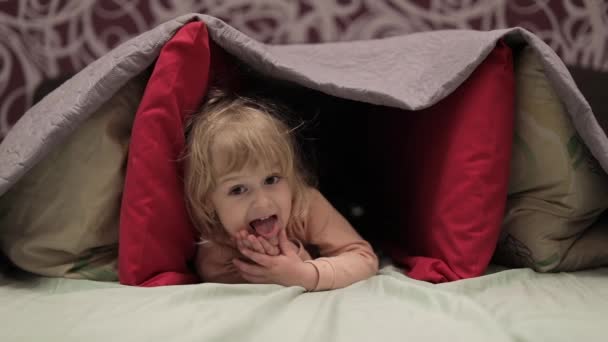 어린 소녀는 침대에 담요와 베개 아래에 숨어있습니다. 은신처의 개념 — 비디오
