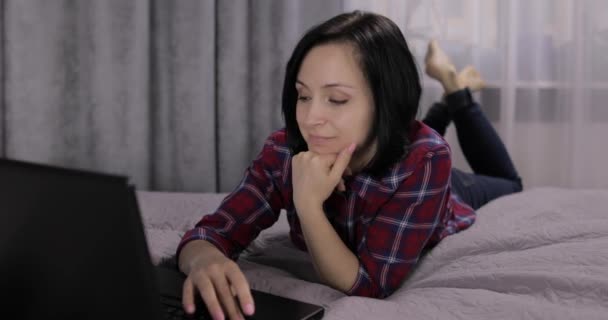 若いブルネットの女性がベッドの上に横たわっていた。チャット、ラップトップコンピュータでの作業 — ストック動画