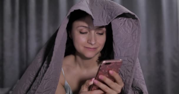 Woman sitting on bed under blanket and enjoying chatting to friend on smartphone — Stock Video