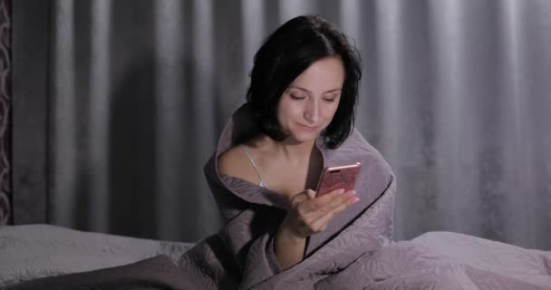 Nő ül az ágyon takaró alatt, és élvezi a csevegést a barátjával okostelefonon — Stock videók