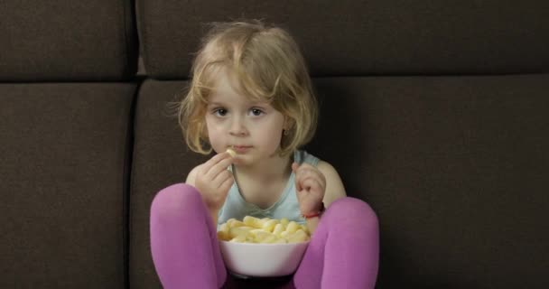Mädchen, das auf dem Sofa sitzt und Windbeutel isst. Kind fernsehen, Papageienkörner probieren — Stockvideo