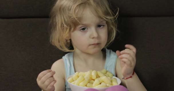 Menina sentada no sofá e comendo sopros de milho. Criança sorrindo e saborear puffcorns — Vídeo de Stock