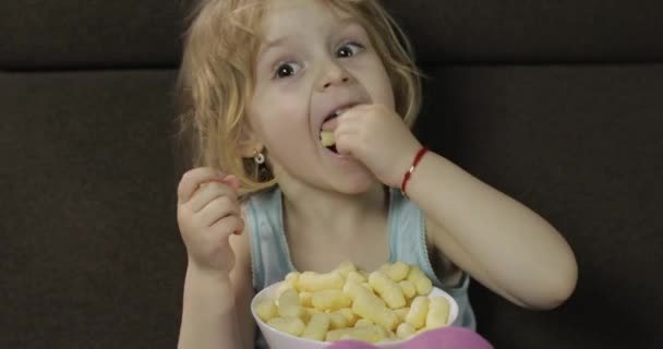 Girl sitting on sofa and eating corn puffs. Child smiling and taste puffcorns — Stock Video