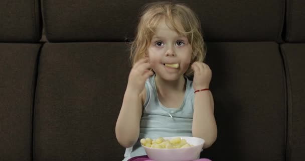 Girl sitting on sofa and eating corn puffs. Child watching tv, taste puffcorns — Stock Video