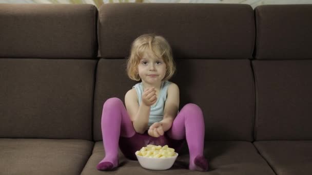 Girl sitting on sofa and eating corn puffs. Child smiling and taste puffcorns — Stock Video