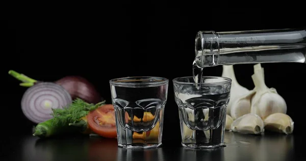 Vierta la bebida alcohólica vodka de la botella en vasos de chupito. Fondo con verduras —  Fotos de Stock