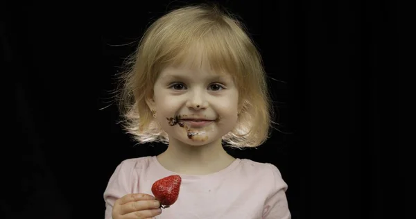 Dítě se špinavou tváří z rozpuštěné čokolády a šlehačkou žere jahodu — Stock fotografie