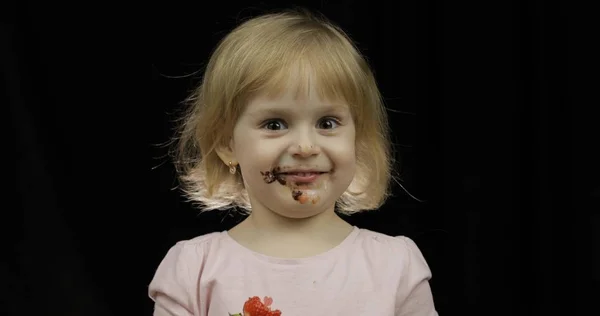 Child with dirty face from melted chocolate and whipped cream eats strawberry