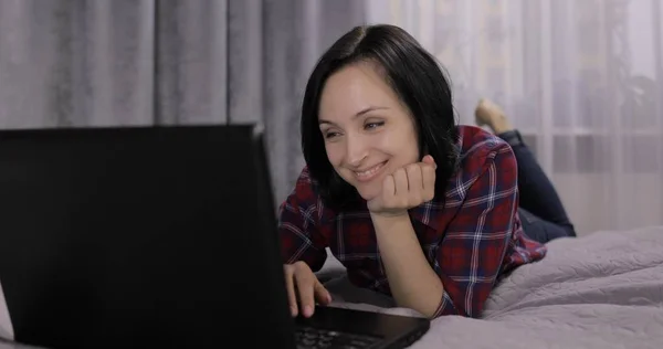 Young brunette woman lying on the bed. Chatting, working on the laptop computer — Stock Photo, Image