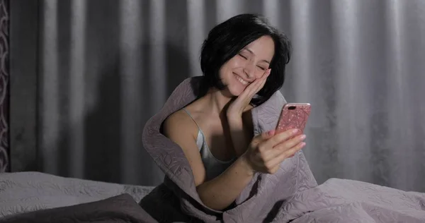 Kobieta siedzi na łóżku pod kocem i rozmawia z przyjacielem na smartfonie — Zdjęcie stockowe