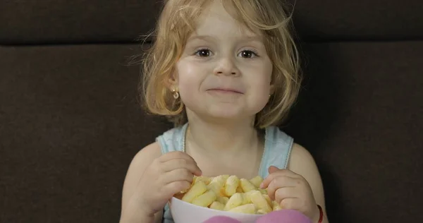 Dívka seděla na pohovce a pojídala kukuřičné pufy. Dětské chuťové koláčky — Stock fotografie