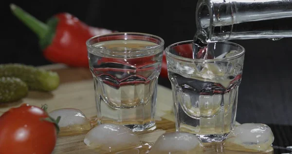 Vierta el vodka de una botella en vasos de chupito con cubitos de hielo sobre la mesa —  Fotos de Stock