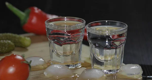 Dos chupitos de vodka en vasos con cubitos de hielo sobre la mesa con verduras —  Fotos de Stock