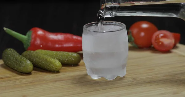 Vierta el vodka de una botella en un vaso helado sobre la mesa con verduras —  Fotos de Stock