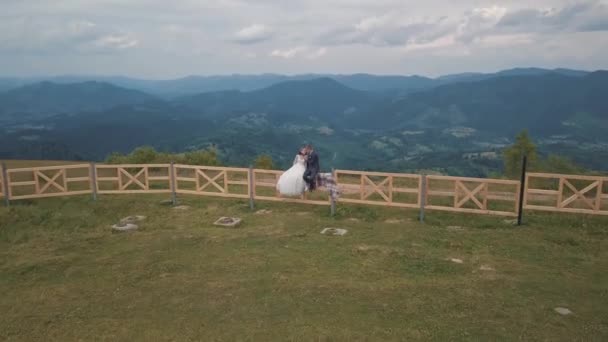 Bräutigam und Braut sitzen auf dem Zaun in der Nähe der Berghügel. Drohnenschuss aus der Luft — Stockvideo