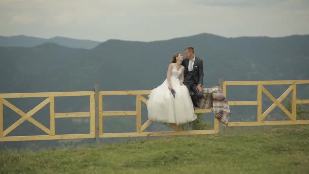 Ženich s nevěstou sedí na plotu u horských kopců. Svatební pár. Rodina — Stock video