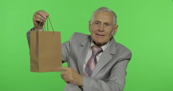 Hombre mayor con bolsa de compras. Mira a la cámara y estira el regalo — Vídeos de Stock
