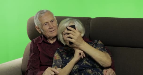 Hombre y mujer ancianos sentados juntos en un sofá y trabajando en un teléfono inteligente — Vídeo de stock