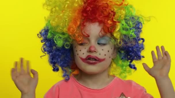 Renkli peruklu küçük kız palyaço şapşal suratlar yapıyor. Eğlenmek, gülümsemek, dans etmek. Cadılar Bayramı — Stok video
