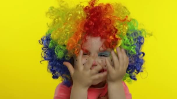 Little child girl clown in colorful wig hides behind her hands and shows funny faces. Halloween — Stock Video