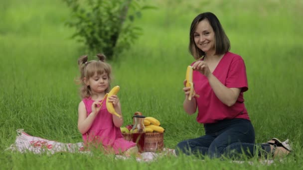 Fin de semana familiar en el picnic. Hija niña con madre en el prado de hierba comer plátanos, divertirse — Vídeo de stock