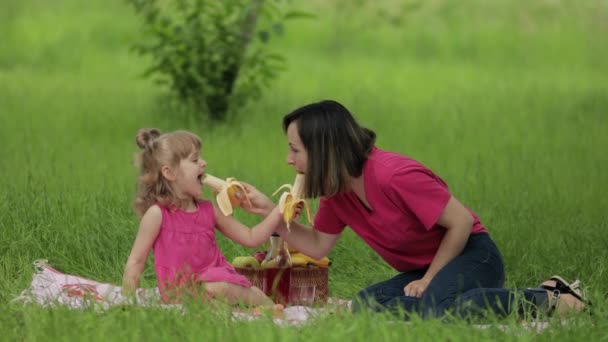 家庭周末野餐。女儿和妈妈一起在草地上吃香蕉，玩得很开心 — 图库视频影像