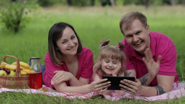 Family weekend picnic. Daughter child girl with mother and father play games on tablet, waving hands — Stock Video