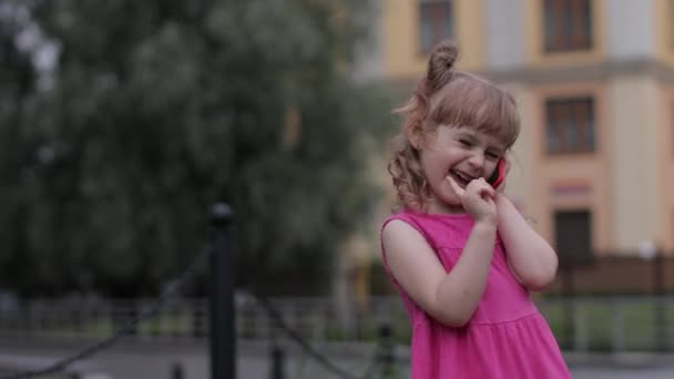 Kind meisje praten met de smartphone buiten. Kind in roze jurk praten door mobiele telefoon op straat — Stockvideo