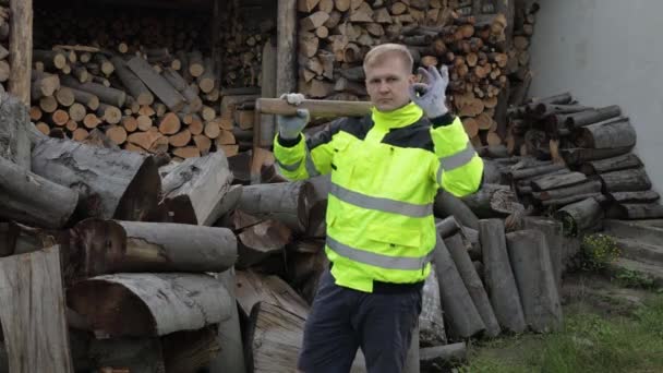 Lumberjack en chaqueta reflectante. Hombre leñador mantenga gran hacha muestran señal ok. Troncos aserrados, leña — Vídeos de Stock