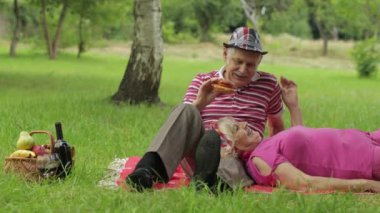 Hafta sonu parkta aile pikniği. Aktif yaşlı beyaz çift battaniyeye oturup hamburger yiyor.