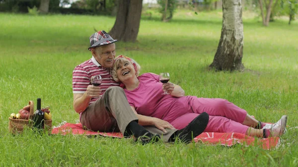 Családi hétvégi piknik a parkban. Aktív idős fehér pár ül a takarón és bort iszik — Stock Fotó