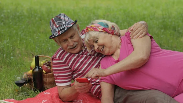 Pique-nique en famille. Vieux grands-parents âgés couple dans le parc en utilisant la navigation en ligne smartphone, chat — Photo