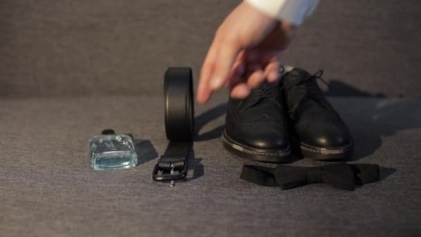El novio toma el cinturón del sofá con perfume, pajarita negra, zapatos. Empresario en la mañana de la boda — Vídeos de Stock
