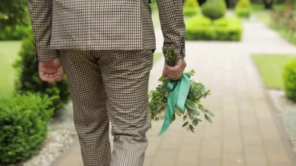 Brudgummen går ner i gränden mellan buskar med en bröllopsbukett till sin älskade brud — Stockvideo